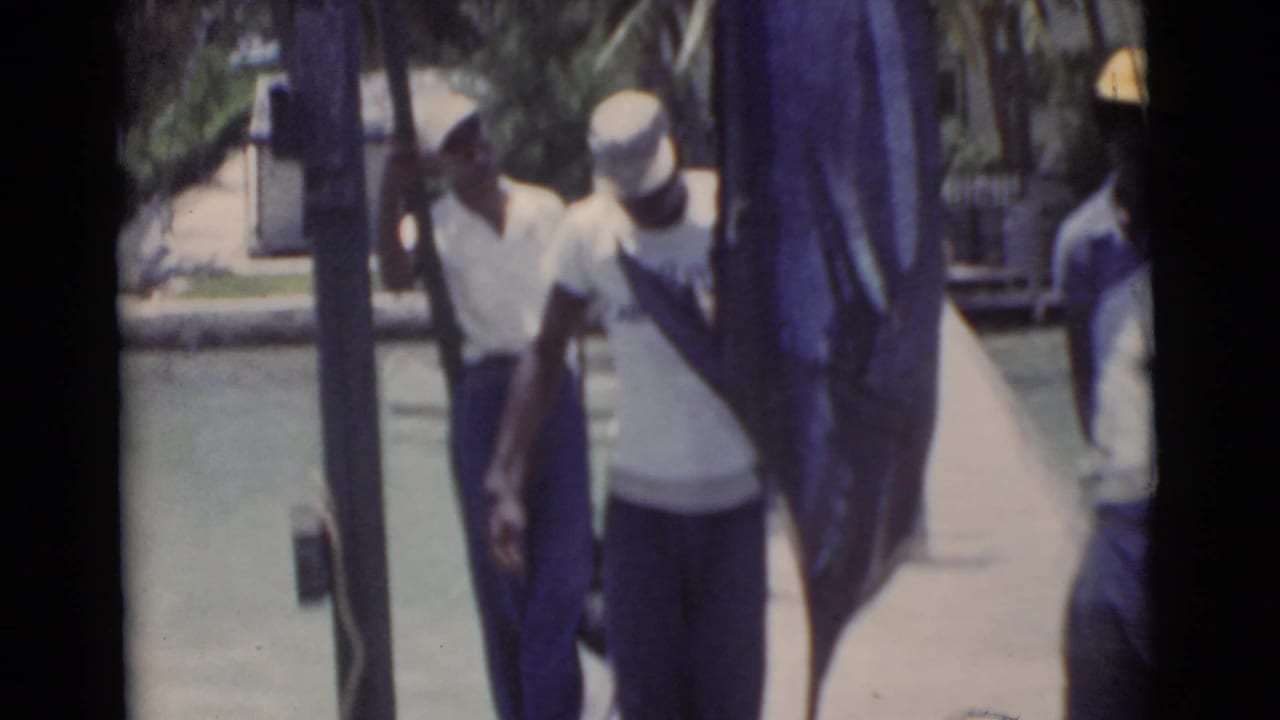Two men steady a large marlin on a dock, with water and palms behind them.