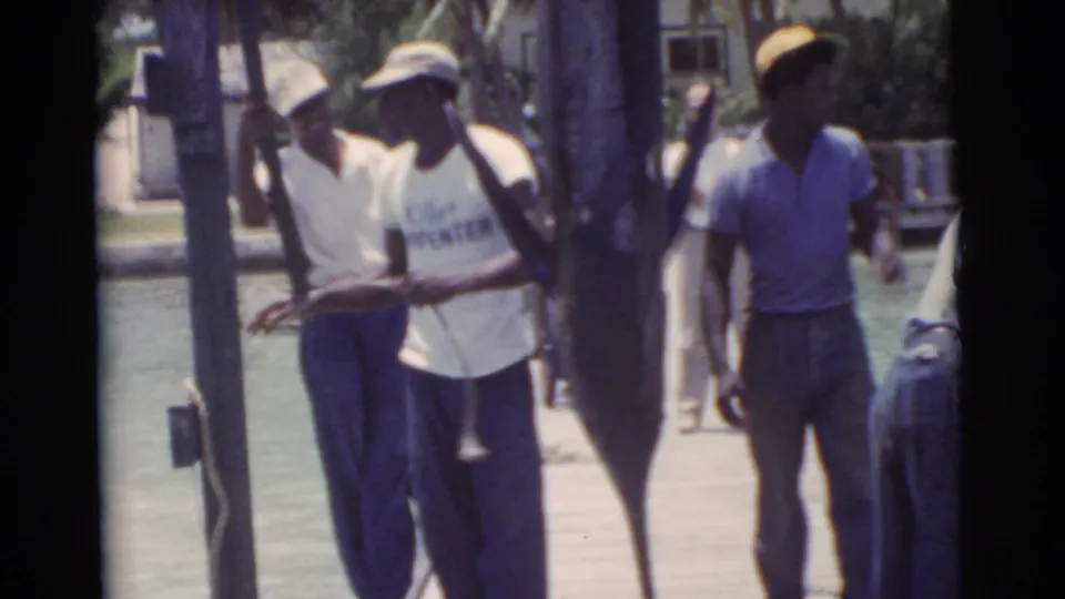 Men in caps and casual shirts stand beside a massive marlin on a Miami dock, with water, palms, and a nearby building behind them.