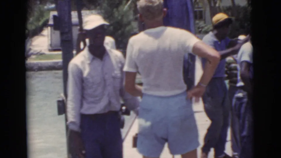 1956 Miami home-movie frame of men gathered on a pier near calm water, with casual summer clothing and caps visible.