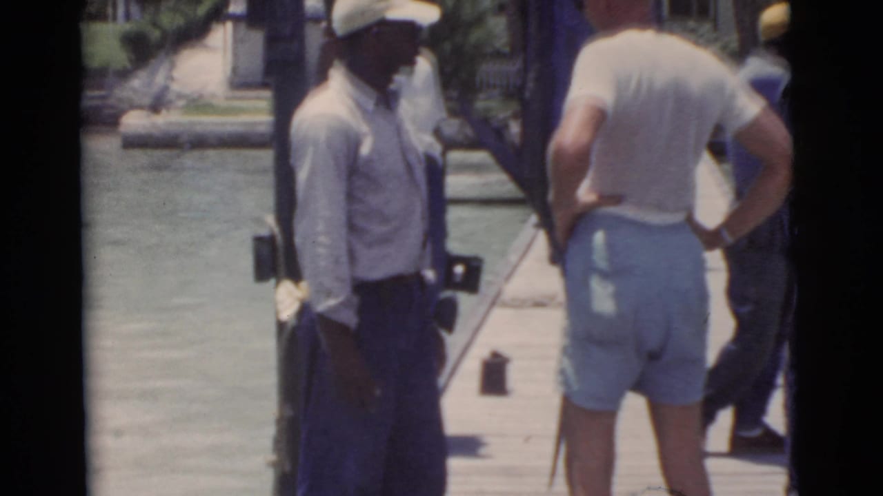 Two men near an upright large fish on a pier beside the water.