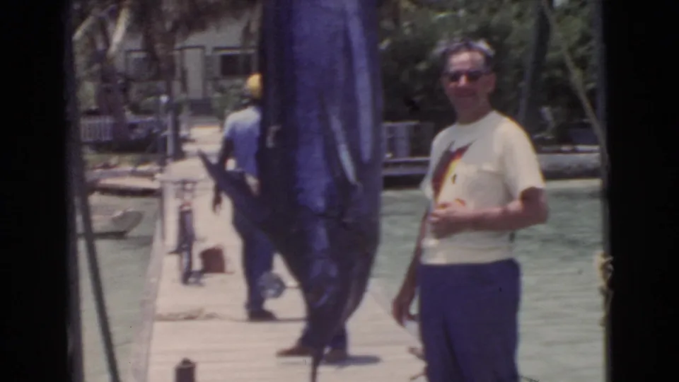 In 1956 Miami, a man in sunglasses poses on a wooden dock next to a large dark blue marlin, with water and palms behind him.