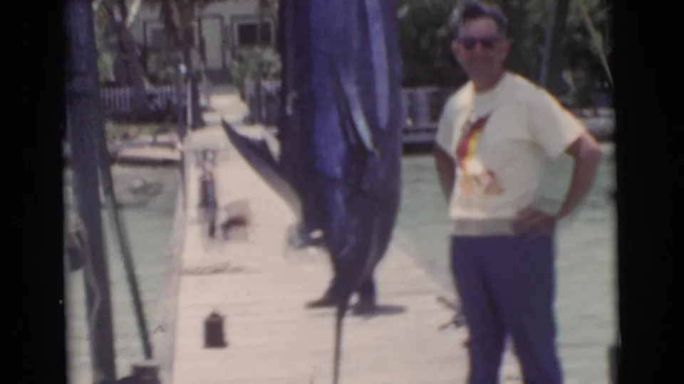 A man in sunglasses and a light shirt stands proudly beside the hanging marlin on a Miami dock, with water and a bicycle nearby.
