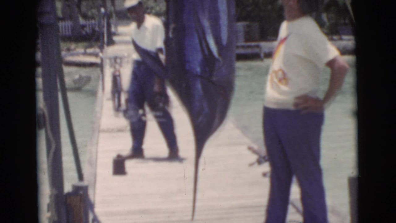 Two men on a dock beside a large hanging swordfish near the water.