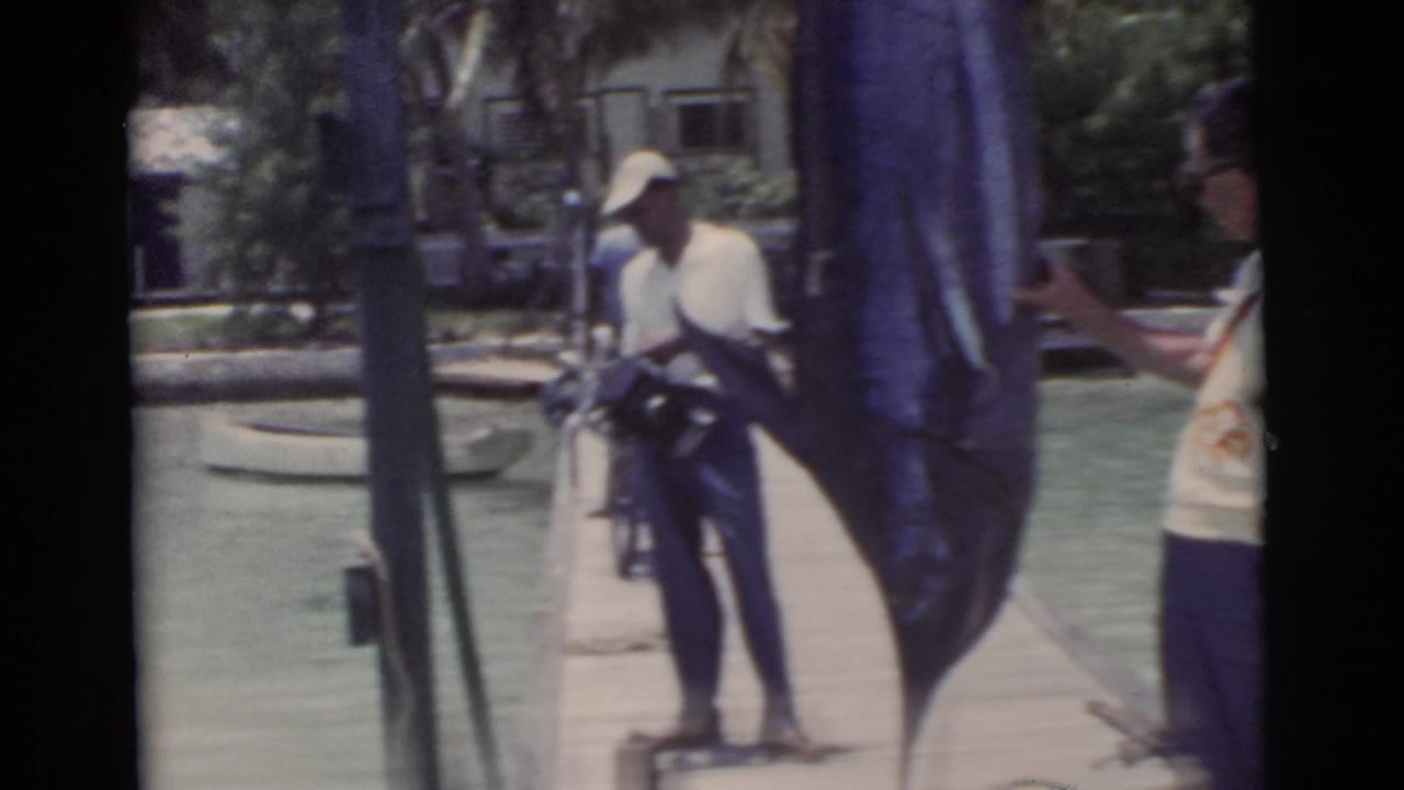 Man in a cap holds a camera near a large hanging swordfish on a dock.