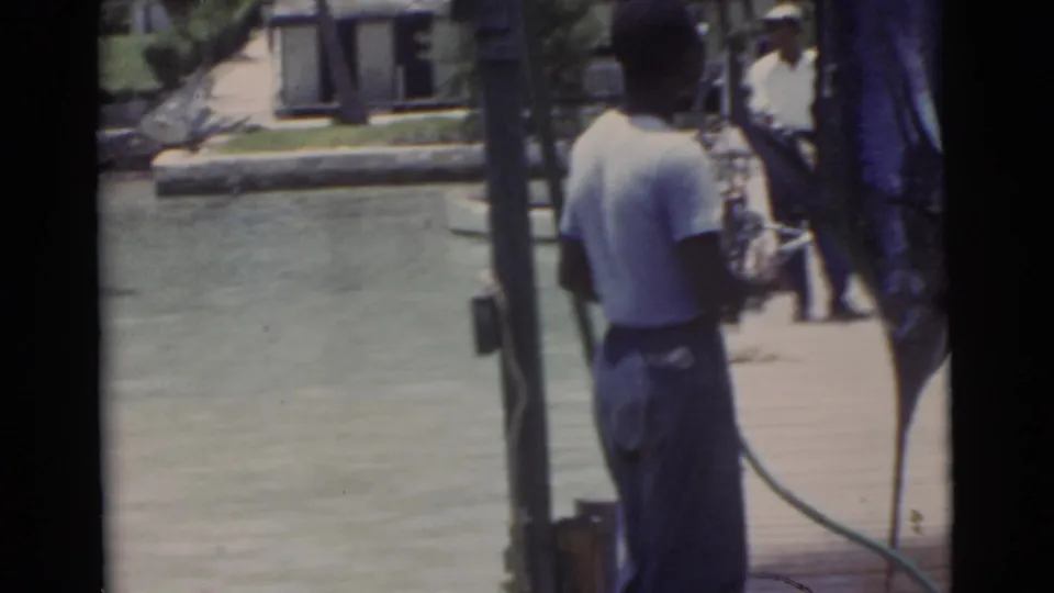 Rear view of an adult man on a Miami dock near a suspended swordfish, with water and low buildings in the background.