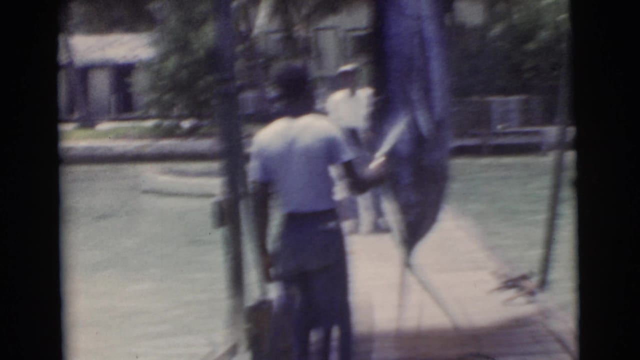 1956 Miami frame of a man in a light shirt and dark pants holding a large fish upright on a narrow wooden dock.