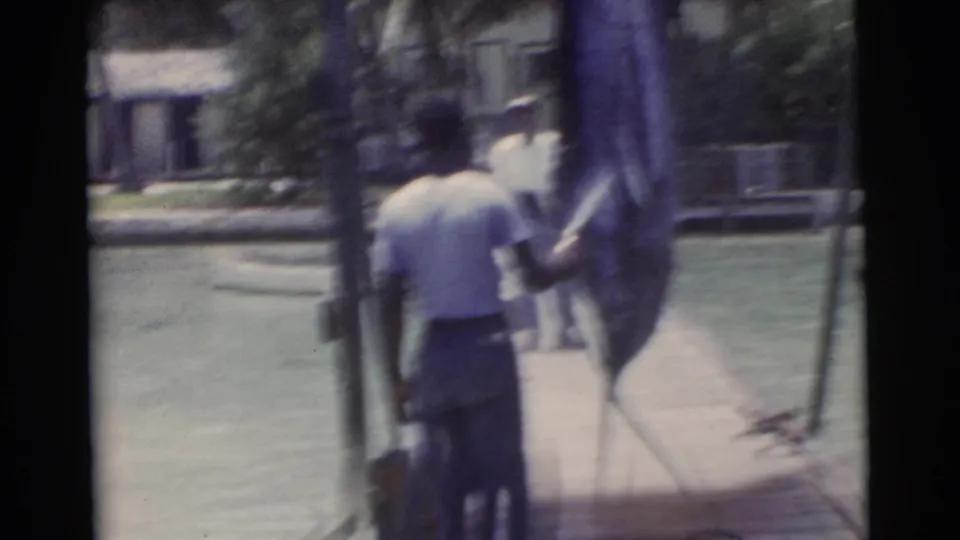 1956 Miami frame of a man in a light shirt and dark pants holding a large fish upright on a narrow wooden dock.