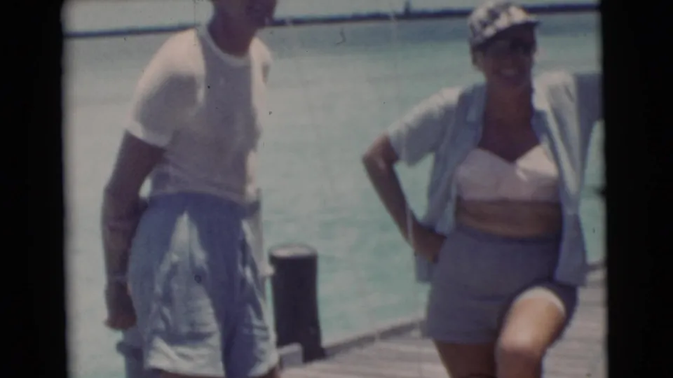 A man and woman in 1950s swimwear pose casually on a Miami dock beside bright turquoise water.