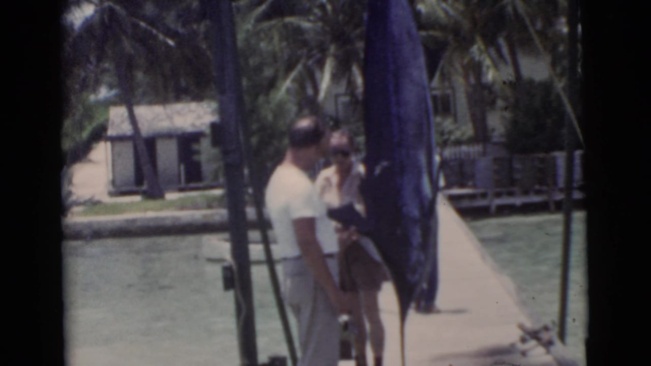 Two men stand on a pier beside a large blue swordfish hanging upright near calm water.