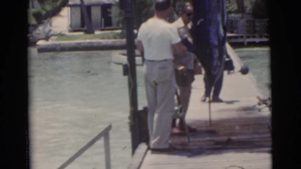 1956 Miami home-movie frame showing a man in light trousers near the suspended swordfish, with another adult partly visible on the dock.