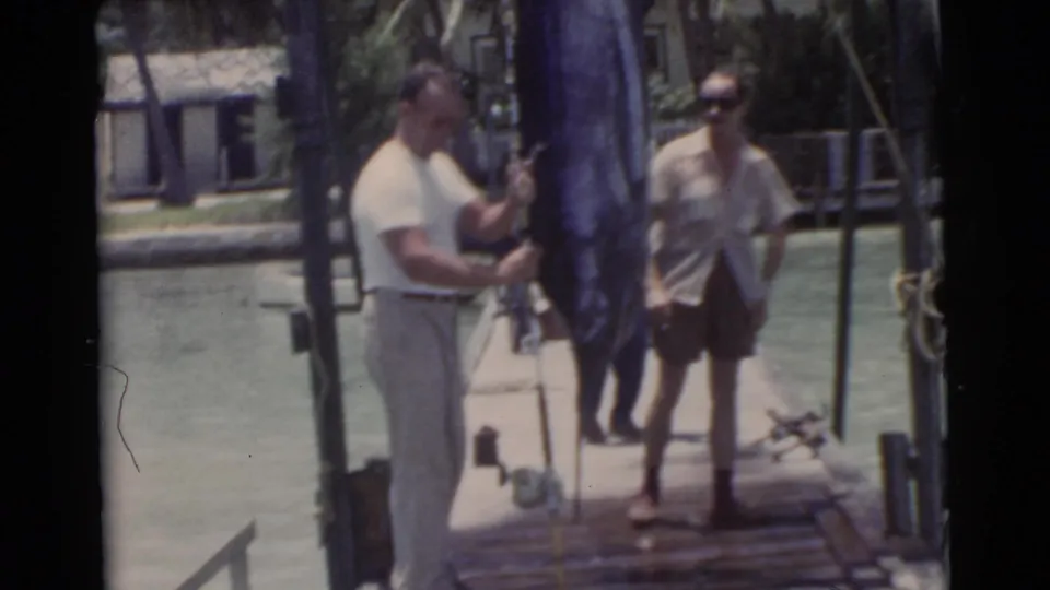 1956 Miami frame showing two adult men on a wooden pier with a large blue swordfish hanging upright between them near calm water and palms.