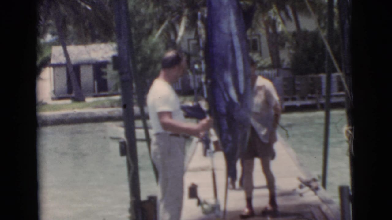 Man on a dock beside a large hanging marlin, with another man nearby and water behind them.