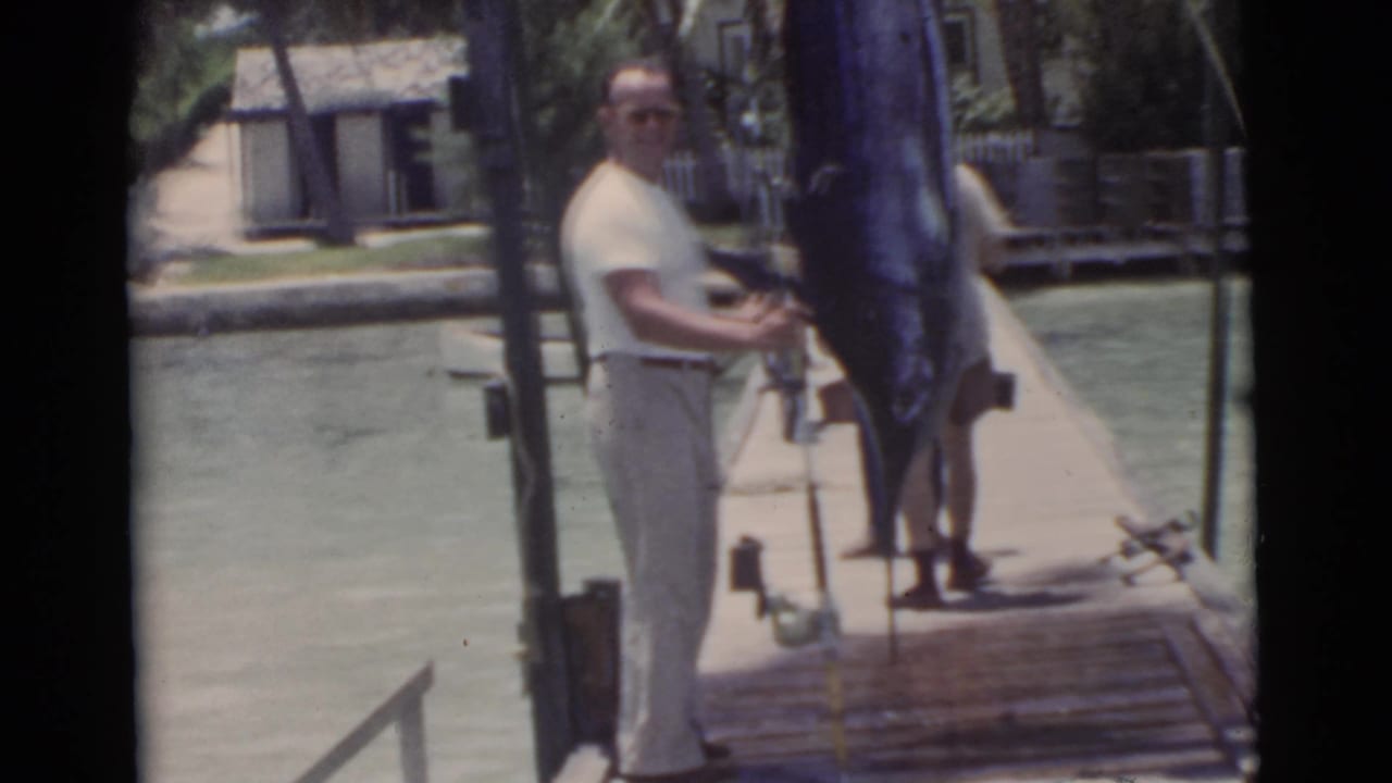 Man in a white shirt stands on a dock next to a vertically hanging marlin over the water.