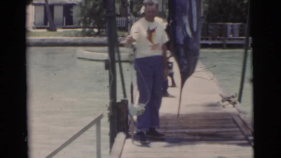 1956 Miami frame showing an adult man facing the camera on a wooden dock beside a large hanging marlin, water, and fishing gear.