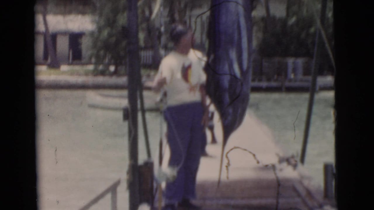 Two men stand on a wooden dock beside a large hanging fish and fishing gear.