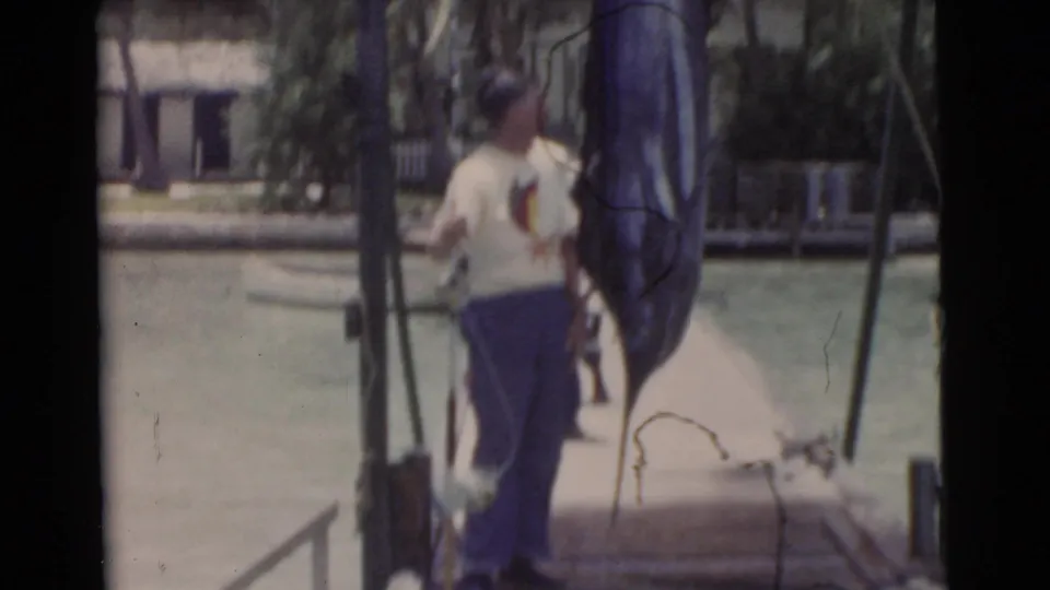 In 1956 Miami home-movie footage, two men stand on a dock near a large marlin with calm water and palms behind them.