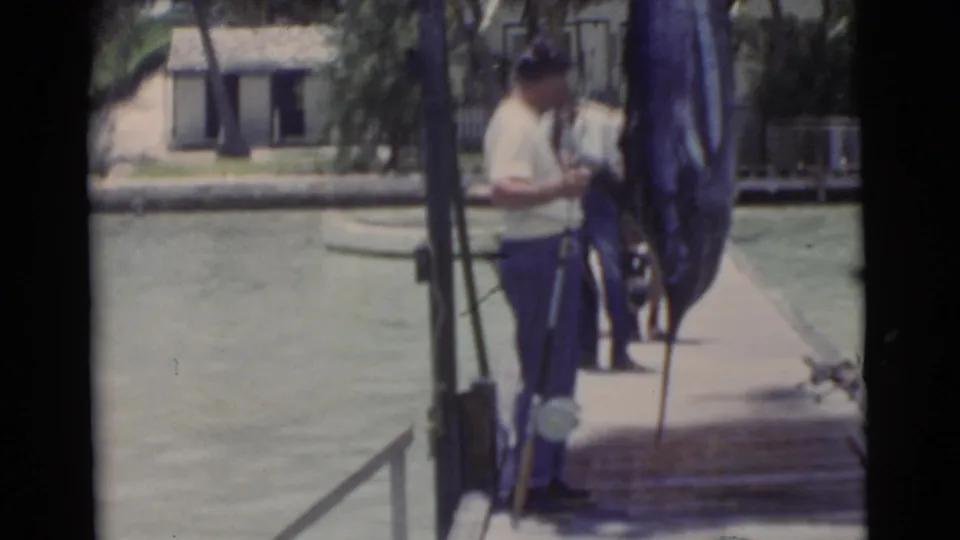 The dockside scene shows a man in a light shirt and blue pants near a hanging marlin, with calm water and palms behind him.
