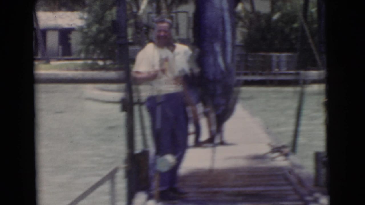 Men standing on a wooden pier beside water, with one figure blurred near the camera.