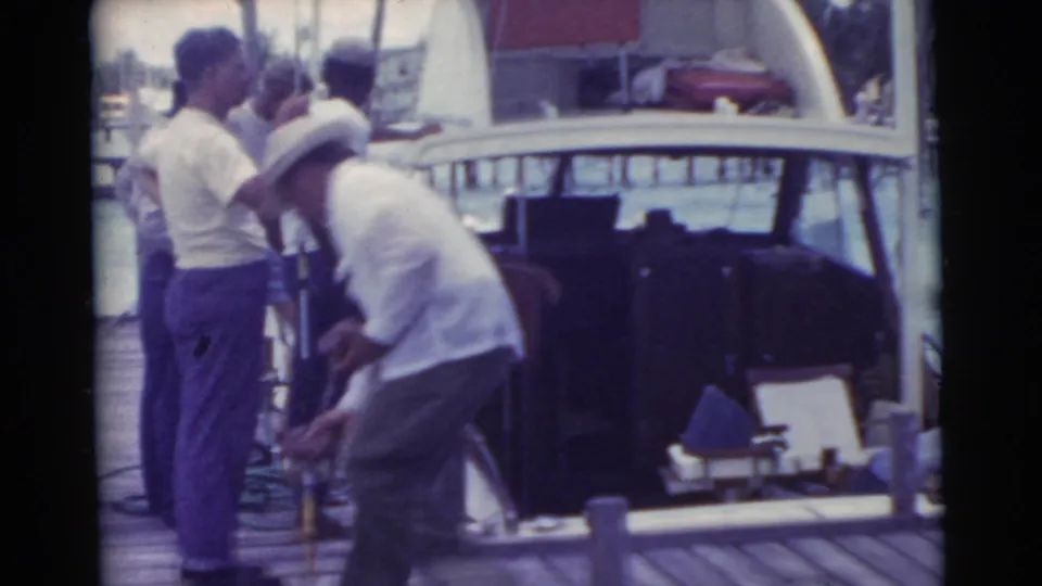 1956 Miami home-movie frame showing men preparing around a docked boat, including one man in a white hat bending near gear.