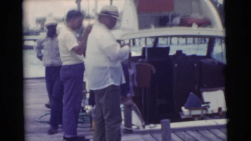 1956 Miami frame of adult men clustered on a wooden dock beside a boat cabin and waterfront railing.