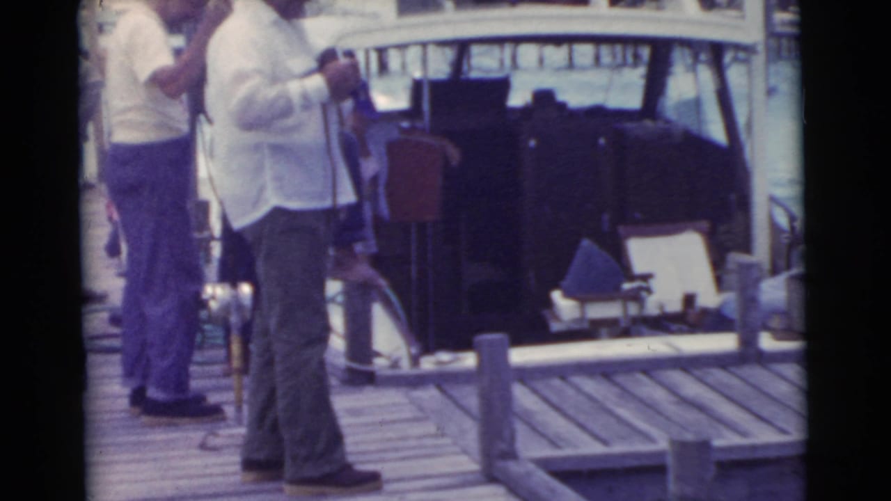 Men in light shirts stand on a wooden dock beside a small boat.