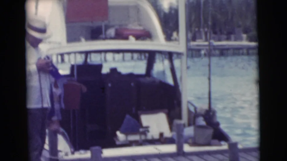 A small boat tied at a Miami dock is seen in soft 1956 8mm color, with adult men partly visible near the left edge.