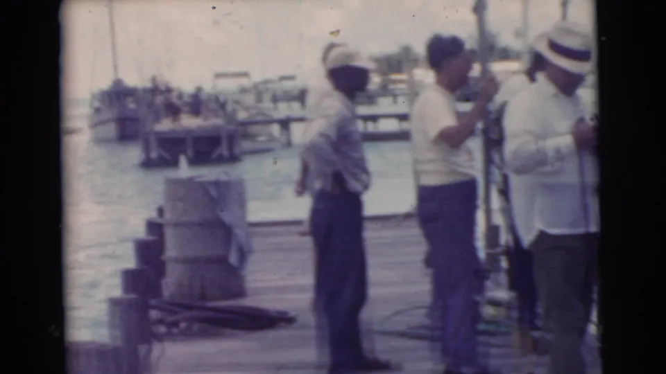 Adult men stand along a Miami dock in 1956 home-movie footage, with harbor water, boats, and dock equipment behind them.