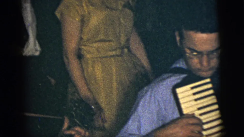 1956 New York City home-movie frame showing a man in a blue shirt playing accordion as a woman in a yellow dress watches in a dim room.