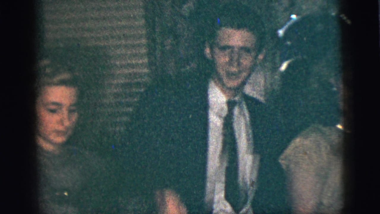 Guests in formal clothing stand together in a dim interior near a microphone.