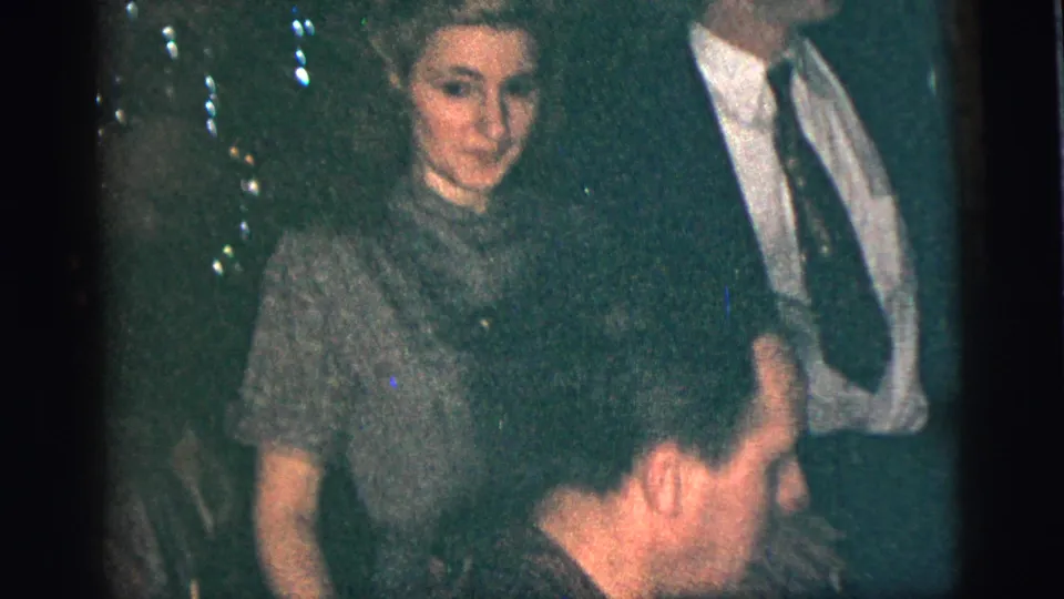 A 1956 New York City home-movie frame shows a woman in a patterned dress, a suited man, and seated guests during an indoor social gathering.