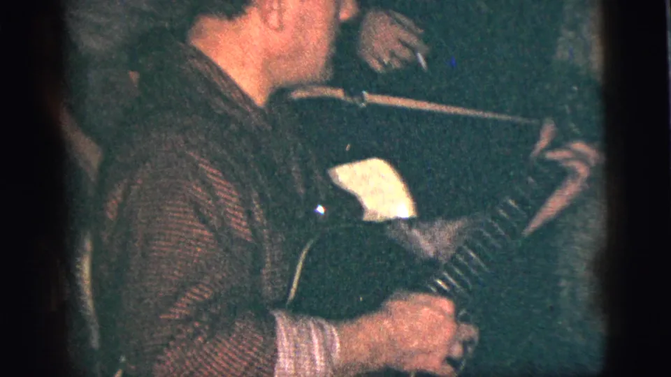 The accordion player is seen in profile near a microphone in the dim private interior, with other guests partly visible around him.