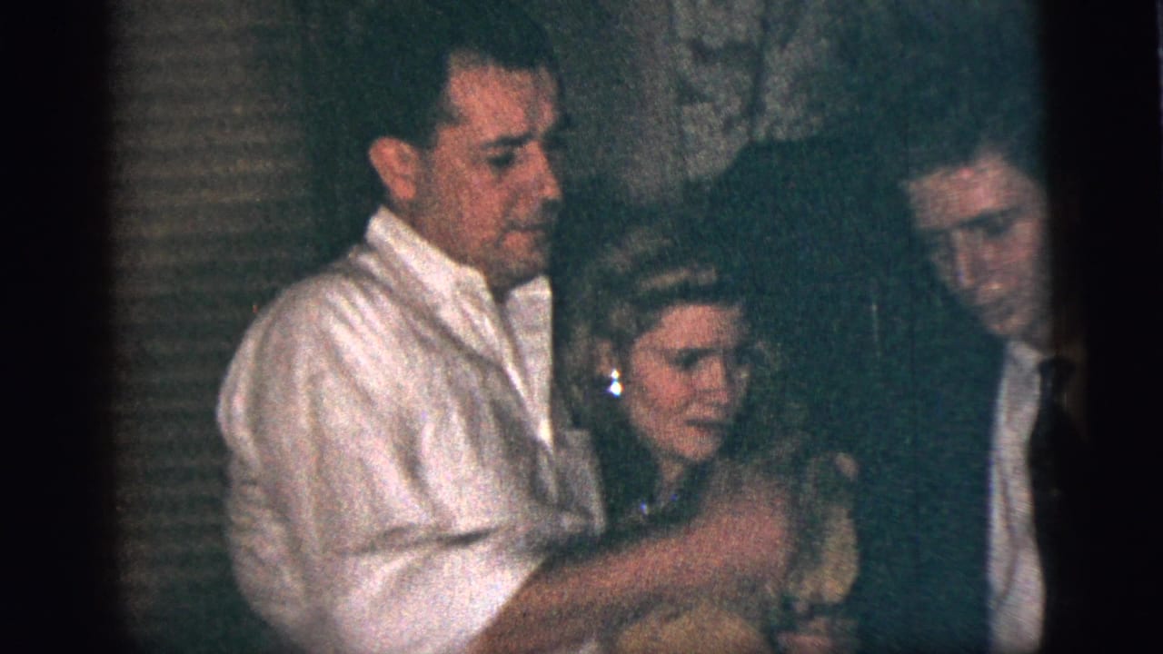 In 1956 New York City home-movie footage, a man in a white shirt stands close to a woman as a young man in a suit looks on.