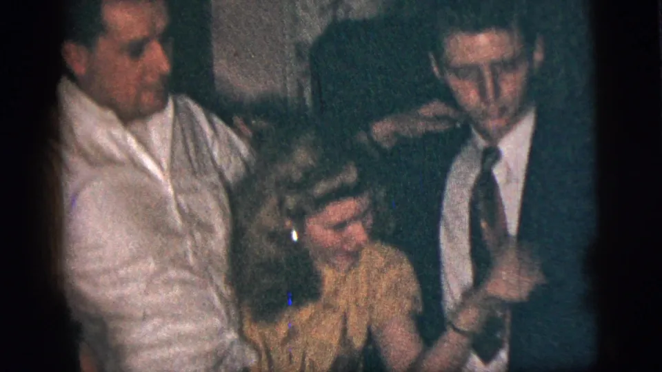 The woman in a yellow dress is framed between the man in a white shirt and the suited young man at the indoor New York City gathering.