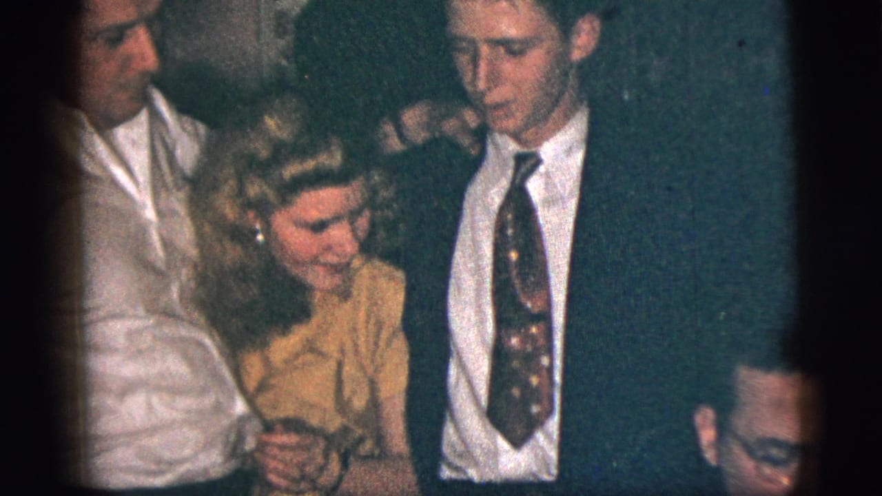 A man in a white shirt and a young man in a dark suit flank the woman in yellow during the 1956 indoor home-movie scene.