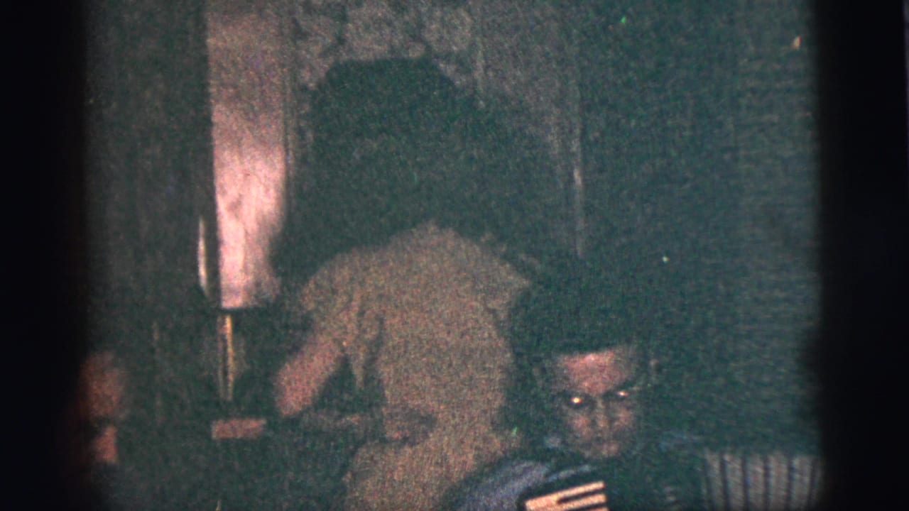 Dim room with people gathered near musicians, including a seated man and a person leaning forward.