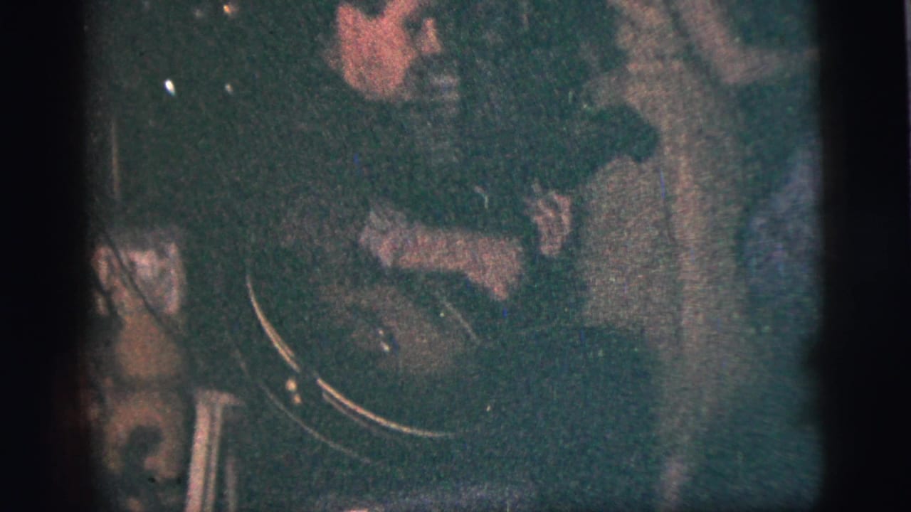 Musician seated in a dark room with a large instrument visible and people nearby.