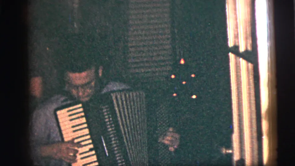 In a 1956 New York City home movie frame, a man in a light shirt plays an accordion near window trim in a dark interior.
