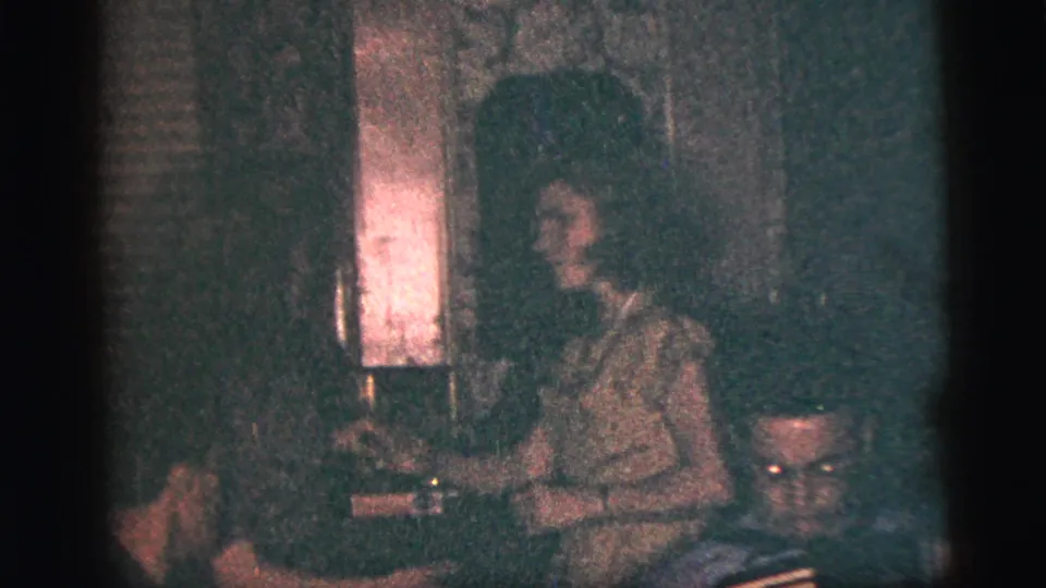 A woman in a dress is seen near another guest in the same dim 1956 New York City home-movie party interior.