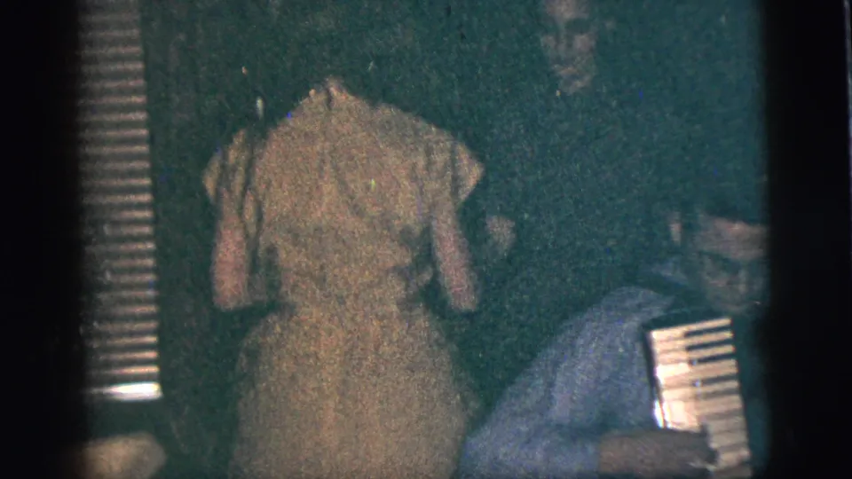 A woman in a light dress appears near seated people in a low-light home interior from the same 1956 footage.