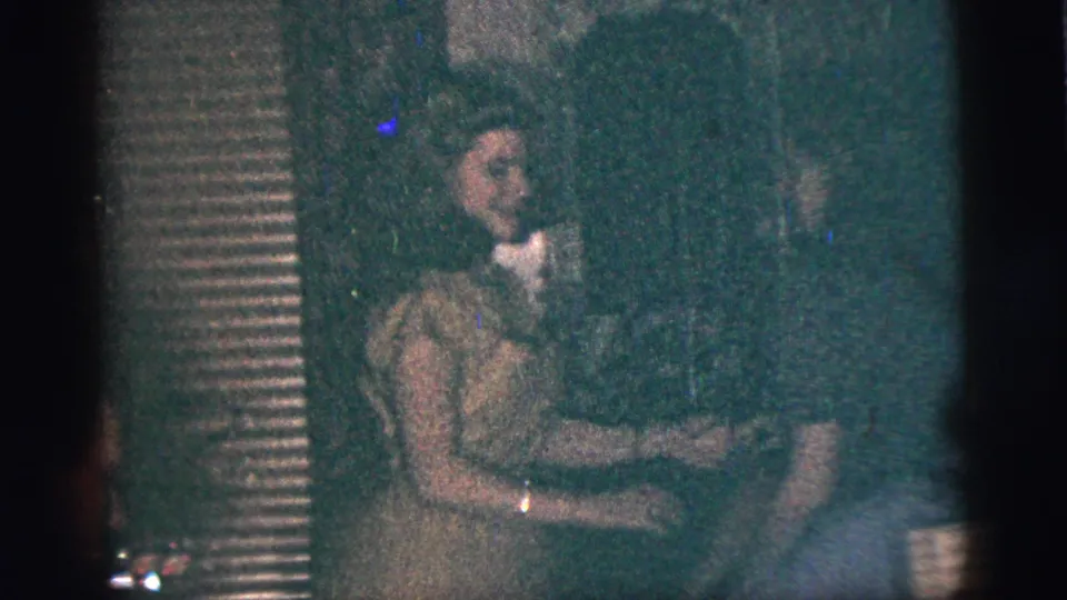 A seated woman and another person appear in a dark home interior, with cups and household details visible.