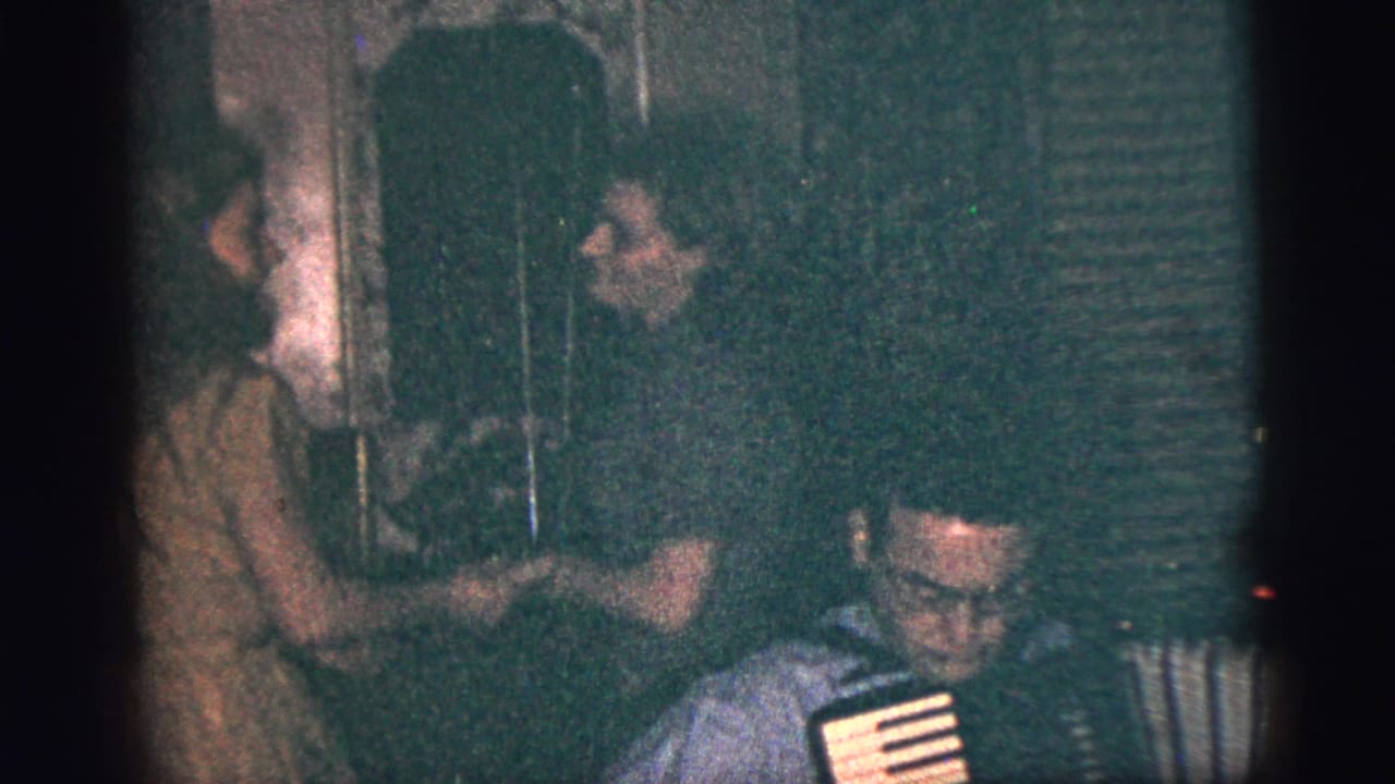 1956 New York City home-movie frame of a seated accordion player with two young women dancing behind him in a private interior.