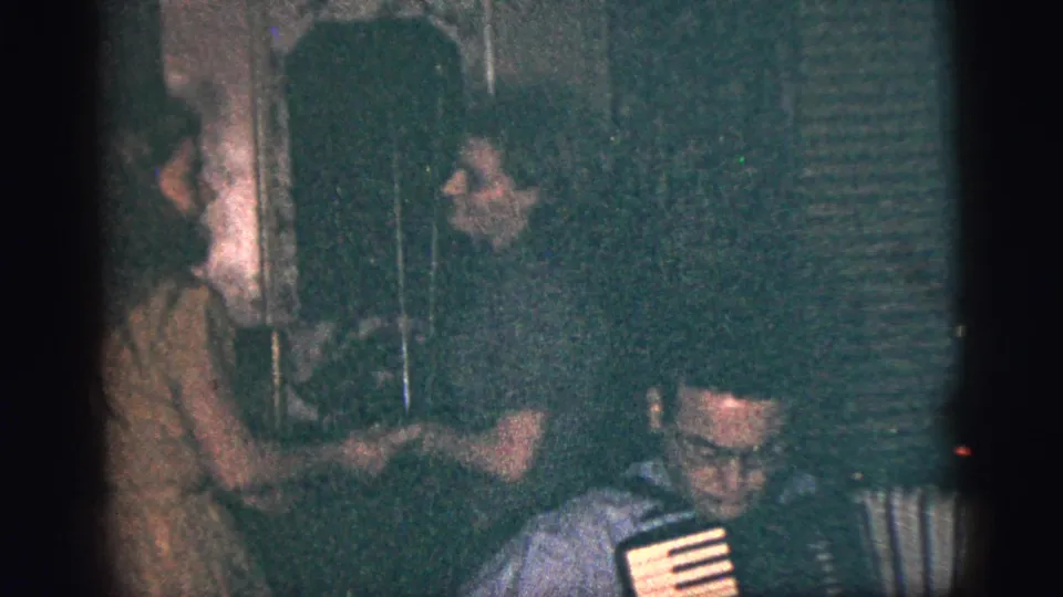 1956 New York City home-movie frame of a seated accordion player with two young women dancing behind him in a private interior.