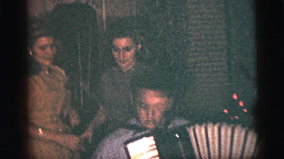 1956 New York City home-movie frame showing adults gathered around a seated accordion player in a private room.