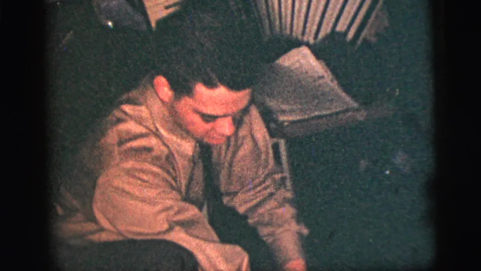 A man in a light shirt leans forward near an accordion and papers in a dim 1956 New York City interior.