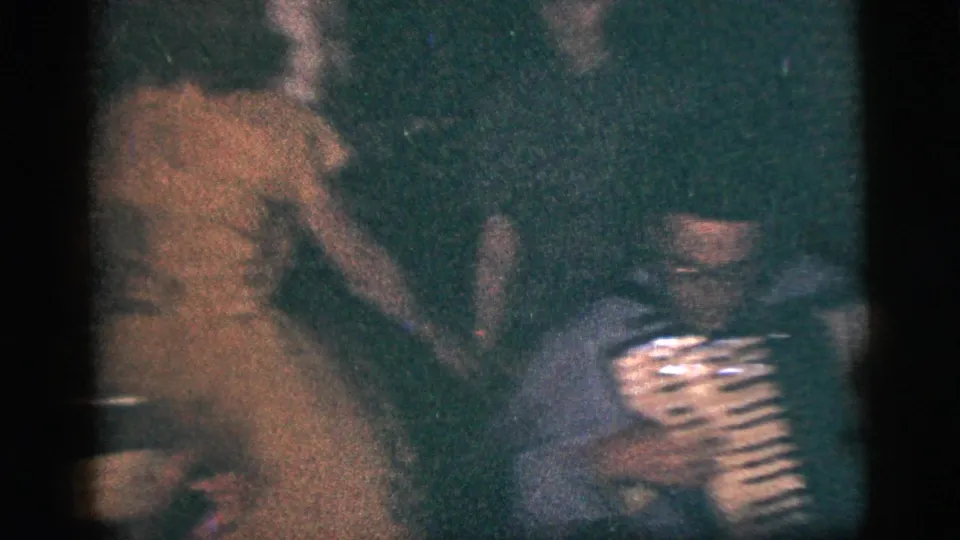 Dim home-movie frame of seated party guests, with an accordion visible in the foreground during the family gathering.