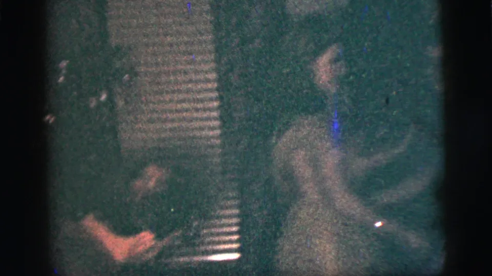A person appears to play an accordion in a dim private interior, with another figure nearby in the home-movie frame.