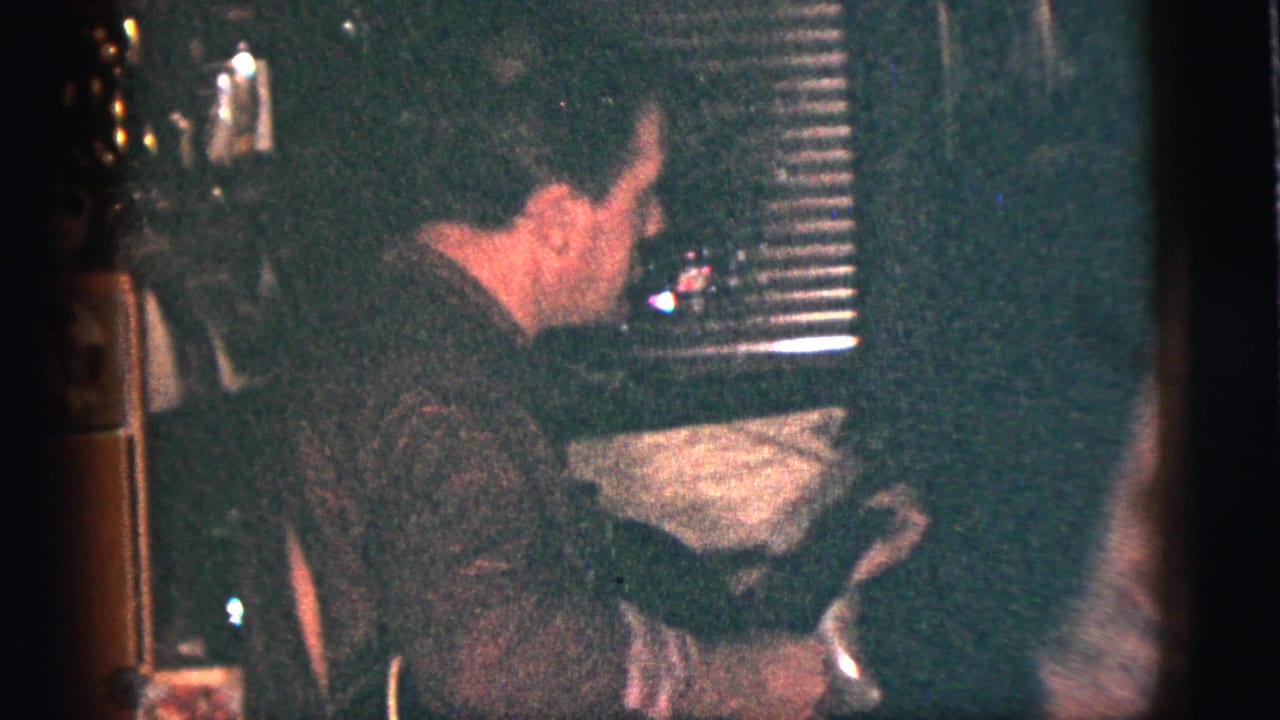 1956 New York City home-movie frame showing shadowed party guests in a dim private interior with blinds and furniture.