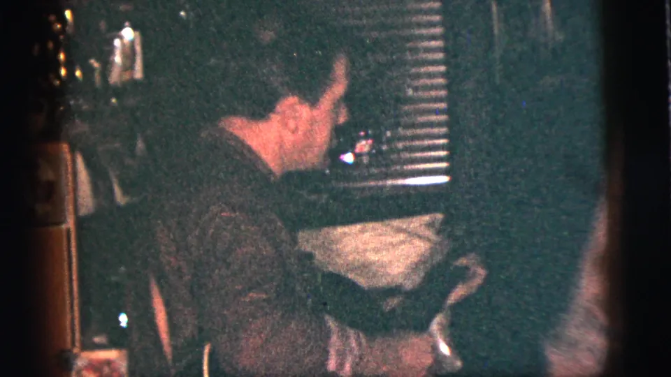 1956 New York City home-movie frame showing shadowed party guests in a dim private interior with blinds and furniture.