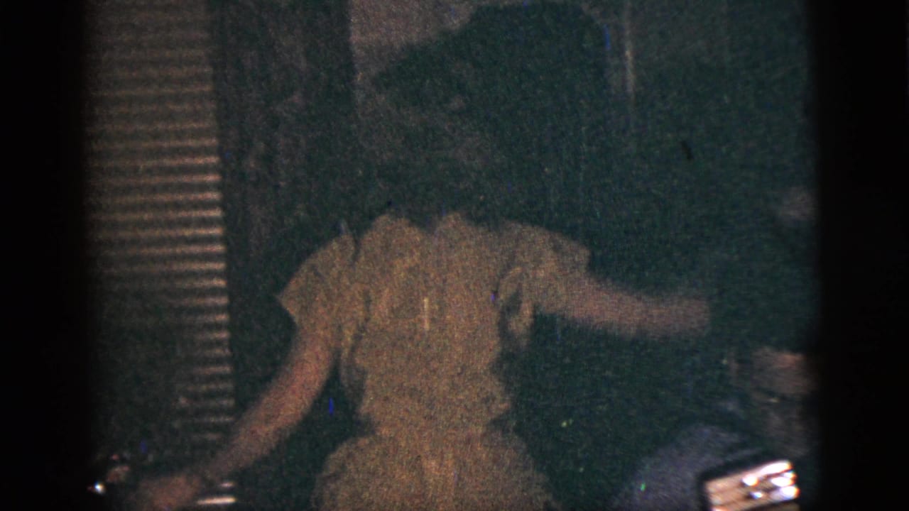 1956 New York City home-movie frame of a woman in a light dress dancing or moving through a dim private room.