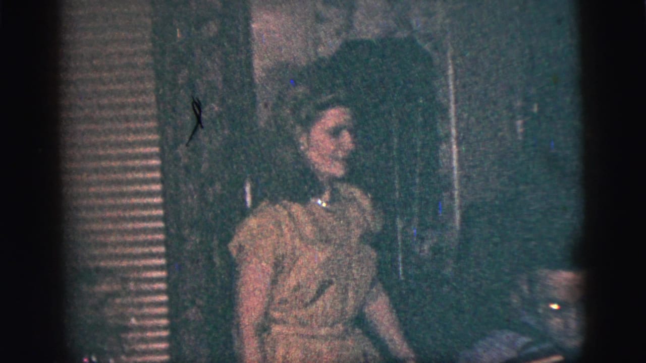 1956 New York City home-movie frame showing a woman in a light dress at an indoor gathering near window blinds.